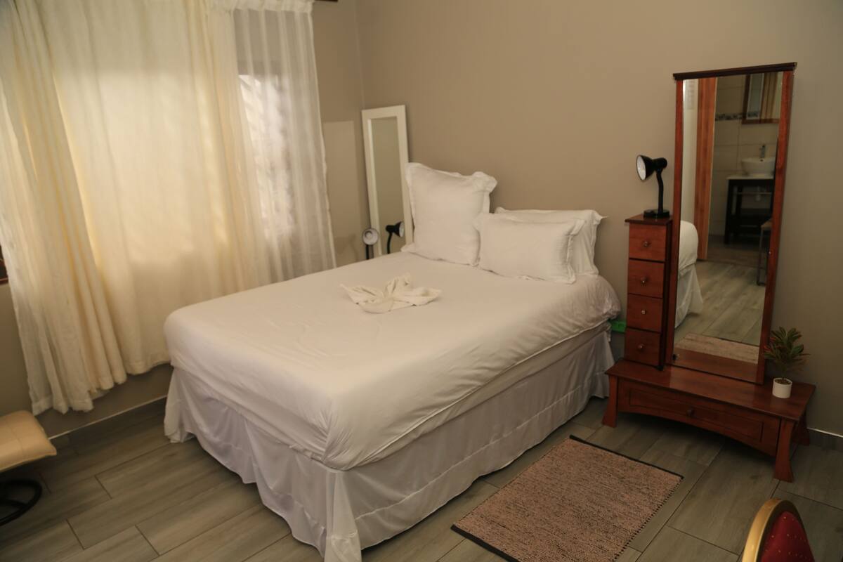 Uma cama bem arrumada com lençóis brancos, um aparador de madeira com um espelho e uma pequena mesa de cabeceira com uma luminária.