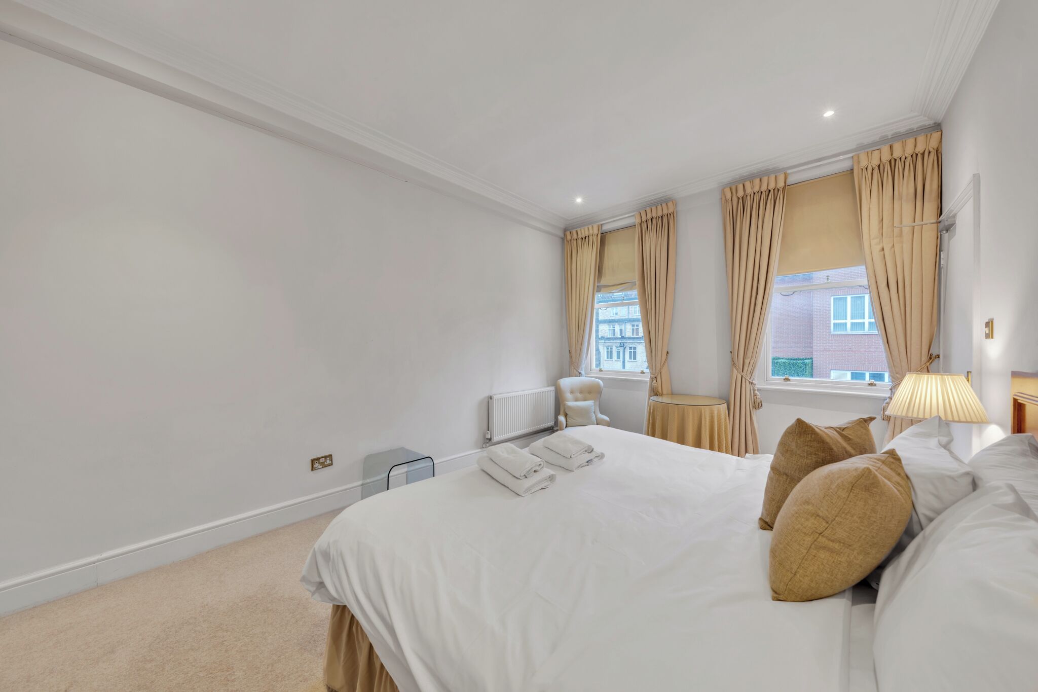 A hotel room with a large bed, beige curtains, a radiator, a chair, and a window with a view of buildings.