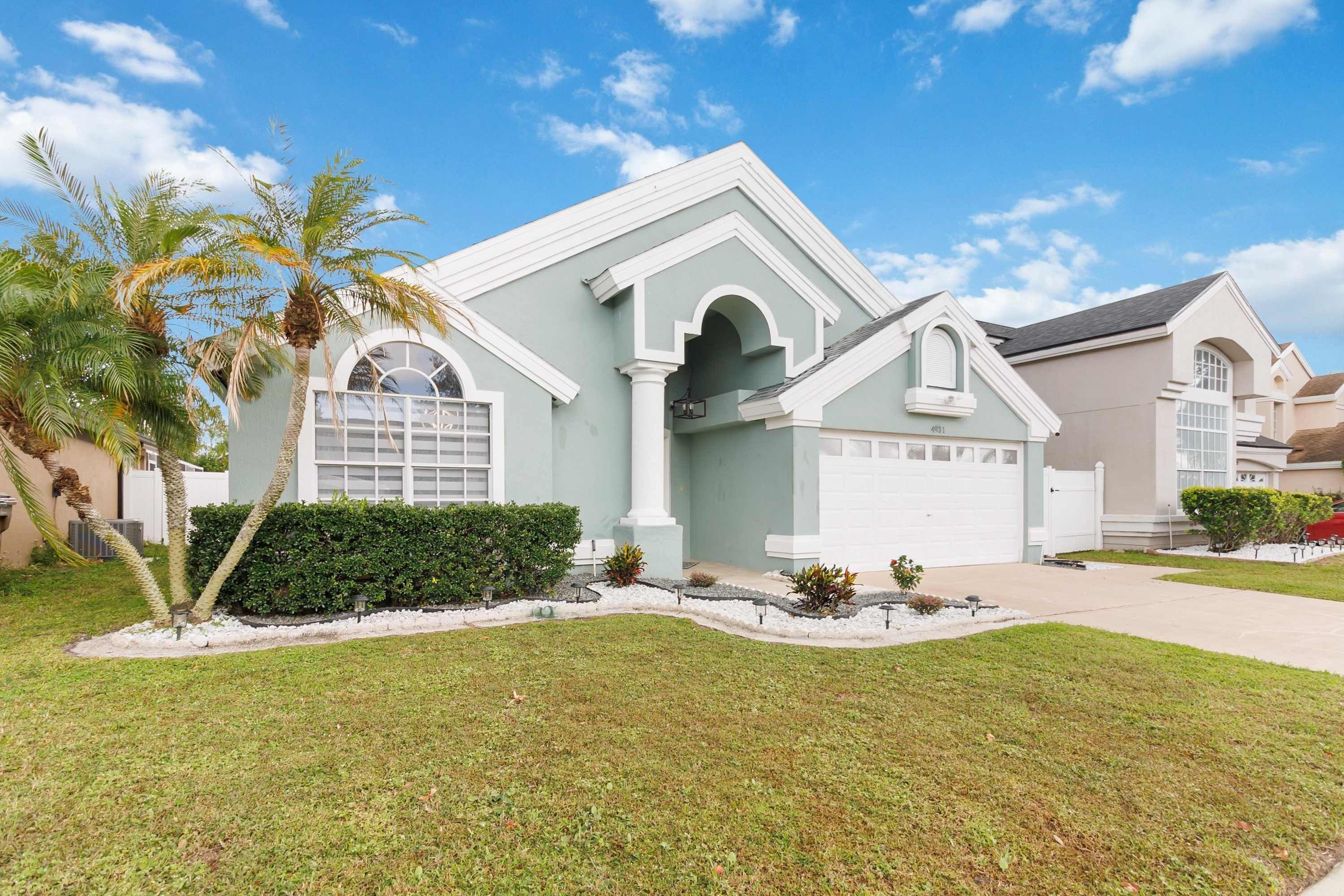 A two-story house with a light green exterior, white trim, and a garage. It has a well-maintained lawn and palm trees.
