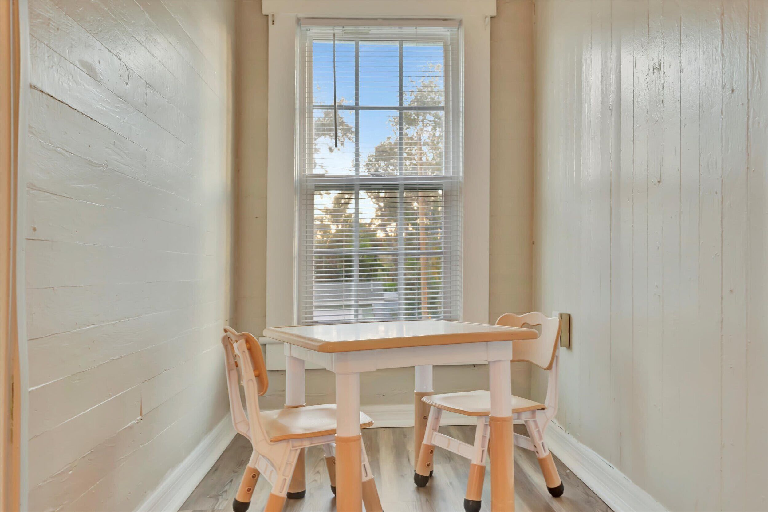 Una habitación de esquina con pared blanca, ventana con persianas, una mesa blanca y dos sillas.