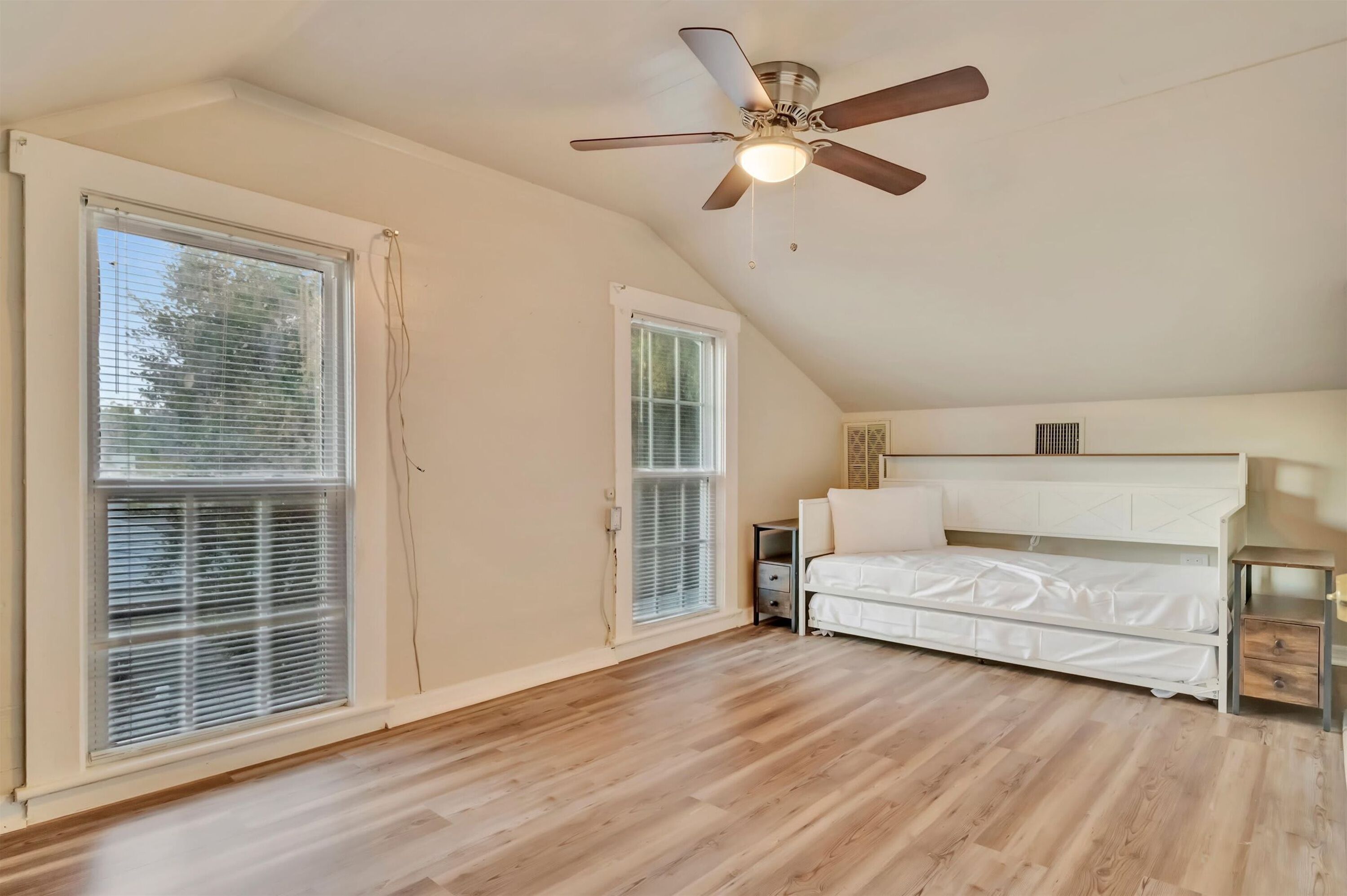 A bedroom with a sloped ceiling, a ceiling fan, a bed, a bedside table, and a window with blinds.