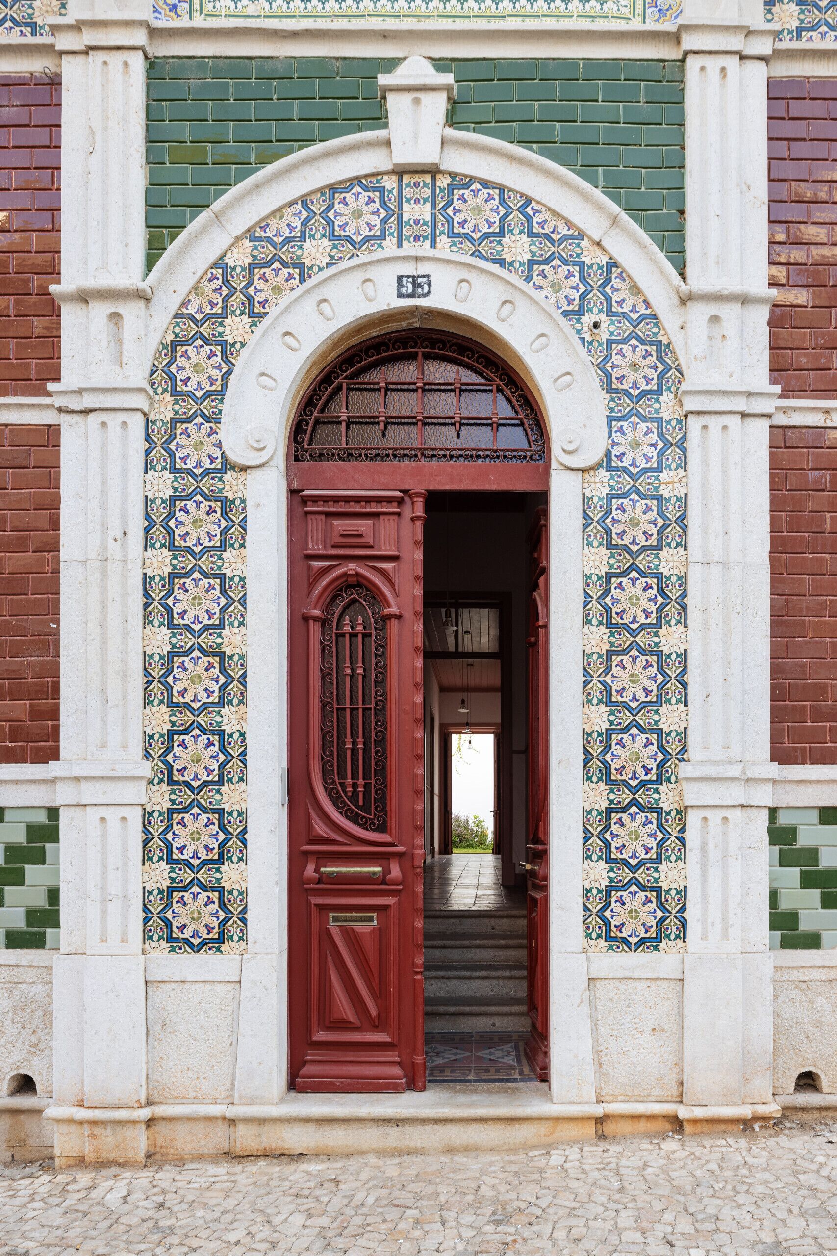 Uma entrada de edifício com porta vermelha, colunas brancas e detalhes em azulejos estampados.