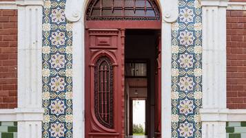 Uma entrada de edifício com porta vermelha, colunas brancas e detalhes em azulejos estampados.