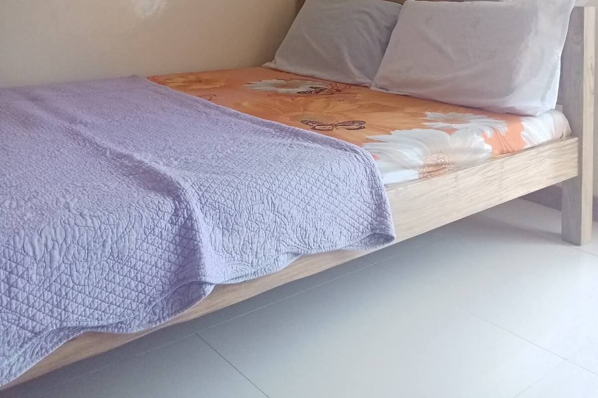 A single bed with a purple blanket, a wooden headboard, and a window with sheer curtains.