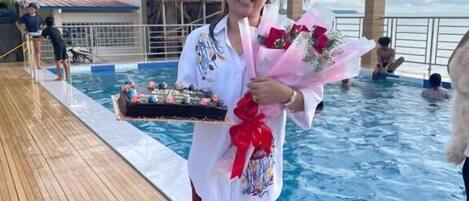 Una persona sostiene una bandeja con comida y flores junto a una piscina, con palmeras al fondo.