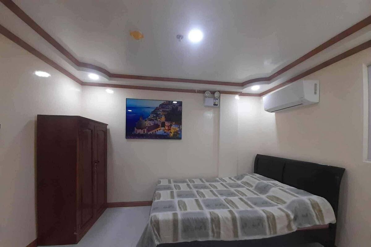 A bedroom with a bed, a wardrobe, a painting, and an air conditioning unit.