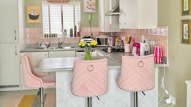Une cuisine avec des meubles blancs, une chaise rose, un comptoir de bar et un luminaire suspendu vert.