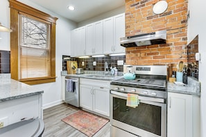 A modern kitchen with white cabinets, a brick accent wall, stainless steel appliances, and a window with blinds. - Walk to High St & Osu! Ultimate Columbus Home Base (Columbus)