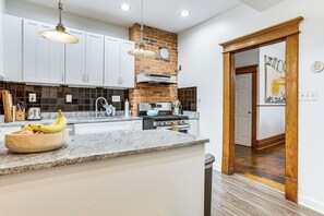 Una cocina moderna con armarios blancos, encimera de granito, pared de ladrillo decorativa y puerta de madera que da a otra habitación.