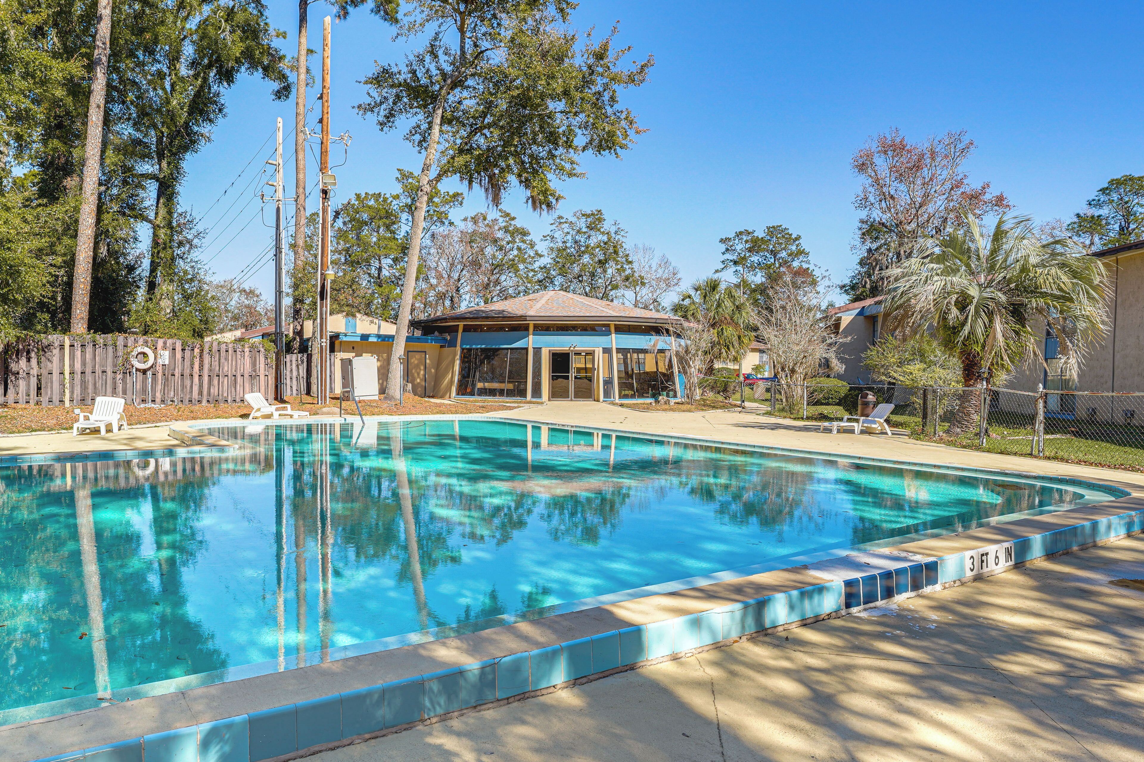 A rectangular swimming pool with a tiled edge, surrounded by a wooden fence, trees, and a covered pavilion.