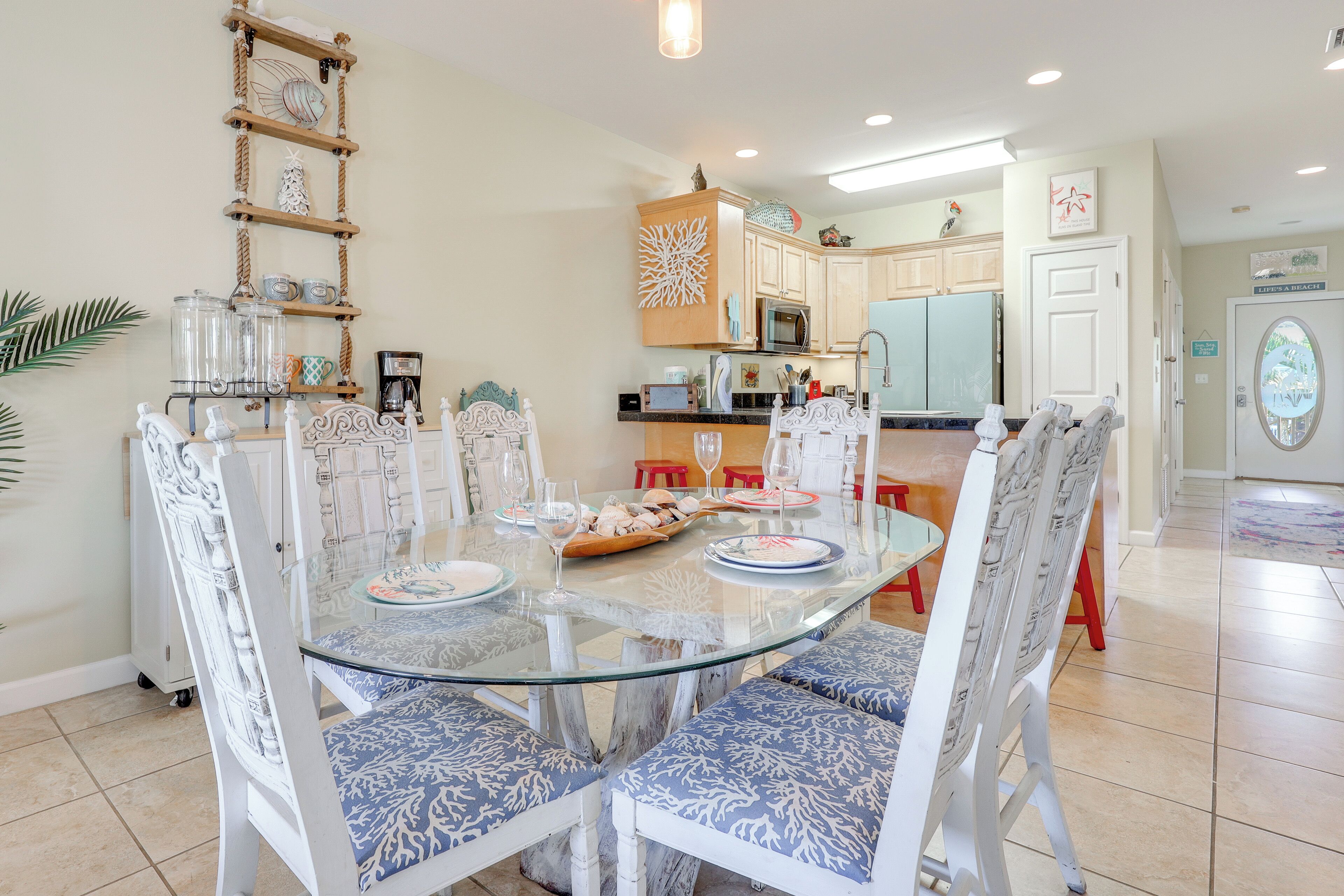 Un coin repas avec une table en verre entourée de chaises blanches avec des coussins bleus, une étagère en bois en forme d’échelle ornée d’objets décoratifs, et un espace cuisine avec des meubles blancs et un four à micro-ondes.