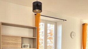 Una habitación con una estantería de madera, una silla negra, un sofá gris, una ventana con cortinas naranjas y un reloj de pared.