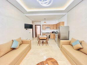 A modern living room with a sofa, a dining table, and a kitchen area with a refrigerator and stove. - New Appart à Guéliz, piscine, balcon et parking (Marrakesh)