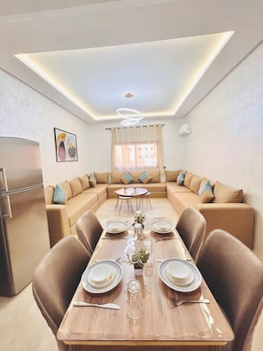 A modern dining area with a long table set for six, surrounded by sofas, a refrigerator, and a ceiling light fixture. - New Appart à Guéliz, piscine, balcon et parking (Marrakesh)