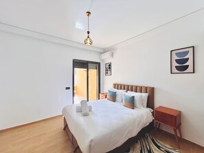 A modern bedroom with a large bed, a wooden floor, a ceiling light, a small wooden nightstand, and a sliding glass door leading to a balcony. - New Appart à Guéliz, piscine, balcon et parking (Marrakesh)