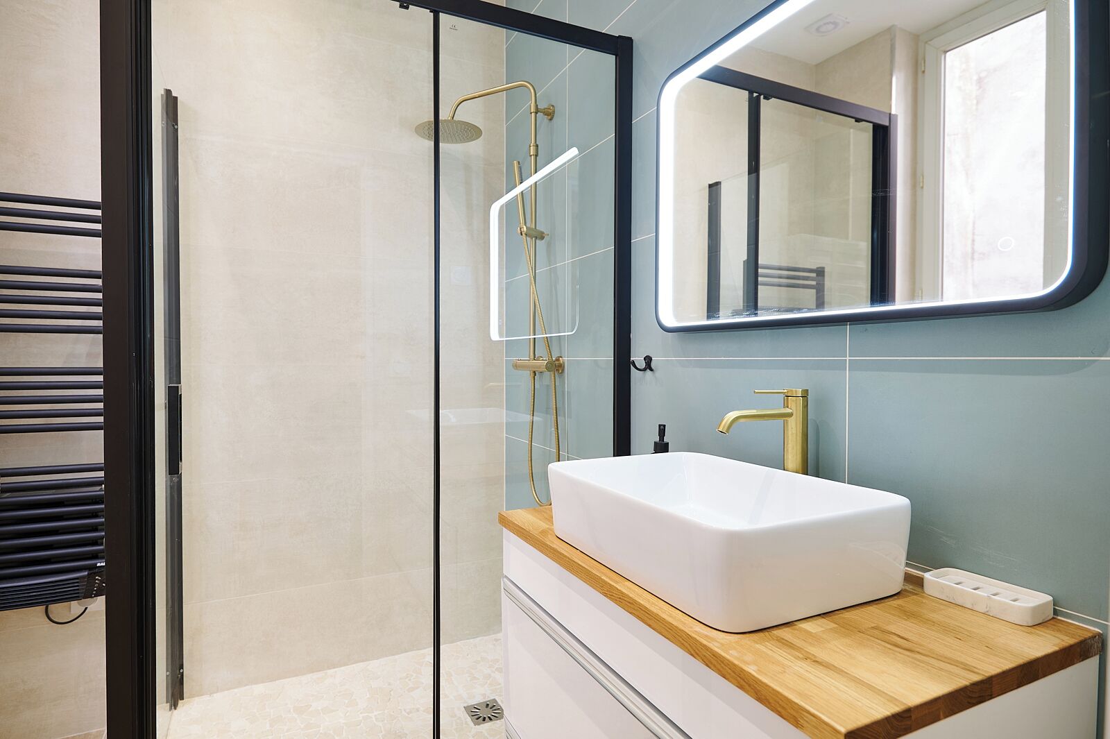 Un baño moderno con ducha envuelta en vidrio, un lavabo blanco con grifo dorado y un mueble de tocador de madera.