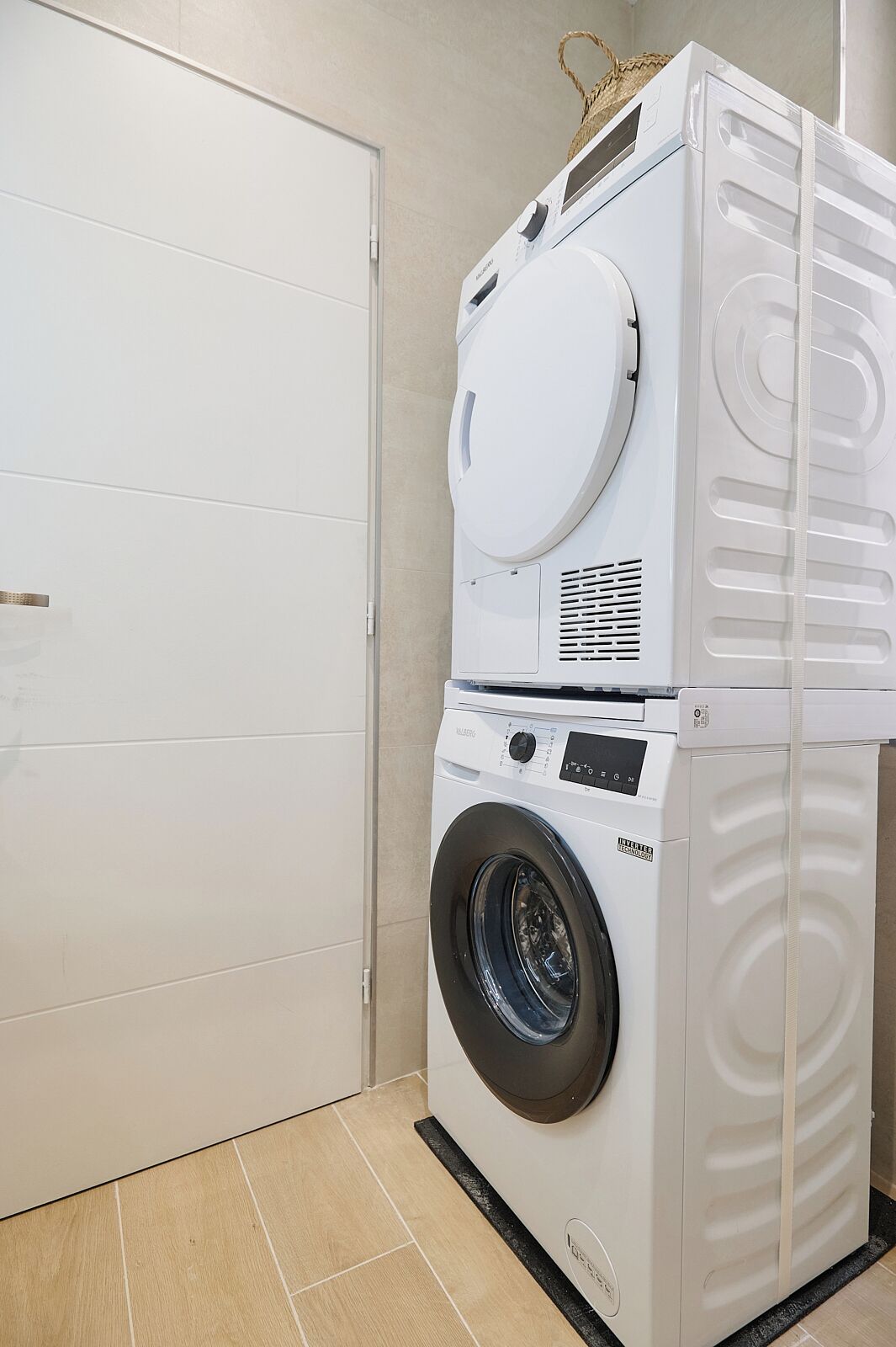 Une machine à laver et un sèche-linge superposés, dans une pièce dont le sol est carrelé et qui est fermée par une porte.