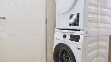 Une machine à laver et un sèche-linge superposés, dans une pièce dont le sol est carrelé et qui est fermée par une porte.