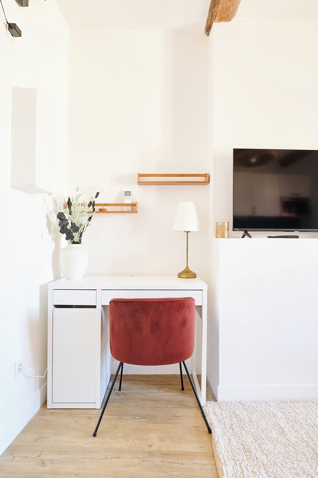 Una habitación moderna y minimalista con un escritorio blanco, una silla roja, un televisor montado en la pared y una planta pequeña sobre el escritorio.