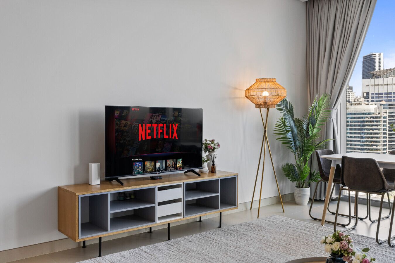 Una sala de estar moderna con un televisor de pantalla plana mostrando el logotipo de Netflix, un mueble de madera para entretenimiento, una lámpara de pie, un comedor con mesa y sillas, y vistas de un paisaje urbano a través de grandes ventanales.