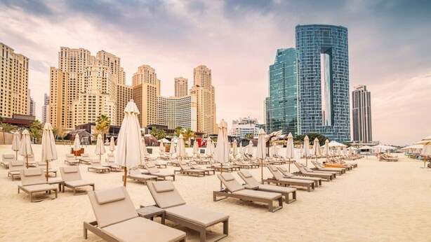 A beach with rows of sun loungers and umbrellas, a modern high-rise building in the background, and a clear sky. - Spacious 2BR in 52|42, Walk to JBR Beach & Marina (Dubai)