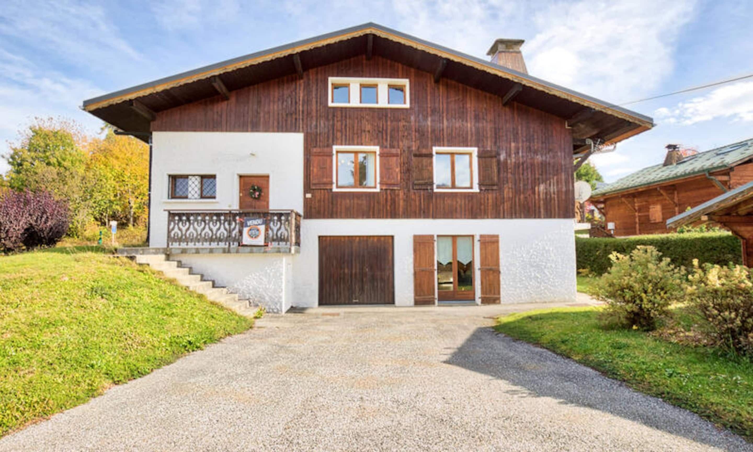 A two-story wooden house with a balcony, a front door, and a gravel driveway.