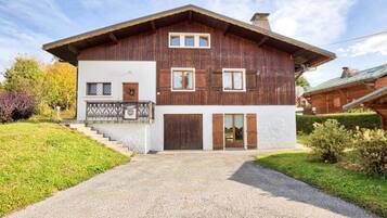 A two-story wooden house with a balcony, a front door, and a gravel driveway.