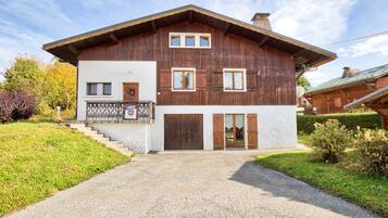 A two-story wooden house with a balcony, a front door, and a gravel driveway.