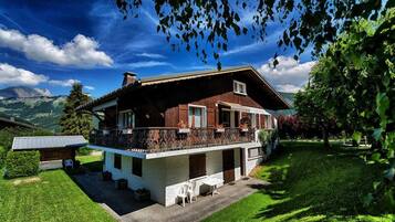 A two-story chalet with a balcony, surrounded by lush greenery and mountains in the background.