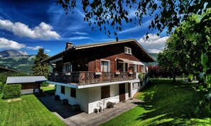 Eine zweistöckige Chalet mit Balkon, umgeben von üppiger Vegetation und Bergen im Hintergrund.