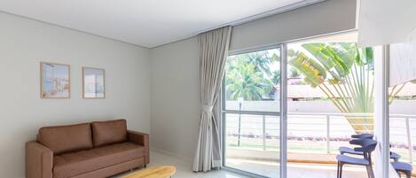 Una sala de estar moderna con un sofá marrón, una mesa de centro de madera y un amplio ventanal corredera que da a un área exterior con sillas y una planta.