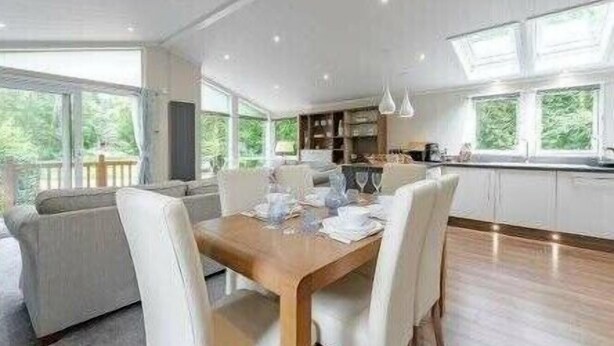 A modern dining area with a wooden table and white chairs, large windows, and a view of greenery outside. - Guelder Rose Lodge - Kelling Heath Holiday Park - 22249 (Holt)