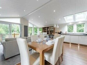 A modern dining area with a wooden table and white chairs, large windows, and a view of greenery outside. - Guelder Rose Lodge - Kelling Heath Holiday Park - 22249 (Holt)