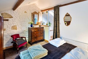 A bedroom with a stone wall, a wooden dresser, a red chair, a bed with white bedding, and a window with curtains. - Little Roost (Lyme Regis)