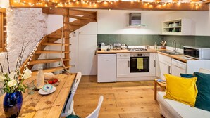 Et hyggeligt køkken med træbjælker, et spiseområde med et bord dækket til to personer, og en trappe til en etage ovenover.