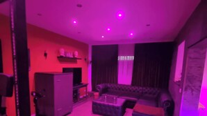 Uma sala de estar com sofá, apoio para os pés e mesa de centro. Há uma televisão em um suporte e um armário. A parede é vermelha e há uma janela com cortinas.