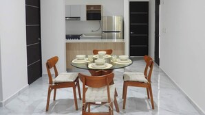 A modern dining area with a glass table, four wooden chairs with cushioned seats, and a kitchen in the background with stainless steel appliances.