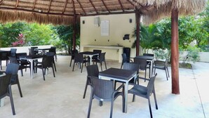 An outdoor patio with thatched roof seating and tables, surrounded by greenery.