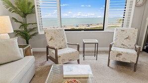 Una sala de estar con vista a la playa, dos sillones estampados, una mesita y una lámpara.