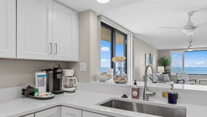 Une cuisine moderne avec des meubles blancs, un évier en acier inoxydable, une machine à café et une vue sur l’océan.