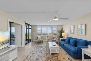 A living room with a blue sofa, white coffee table, and armchairs, a television, and a ceiling fan. - Escape to Loggerhead 511, a direct Gulf-front corner condo on Sanibel Island. This 2-bedroom, 2-bath retreat sleeps 6 and features two private lanais with walkout to beach access, heated pool, pickleball, tennis, comp Golf, and stunning Gulf views. (Sanibel Island)