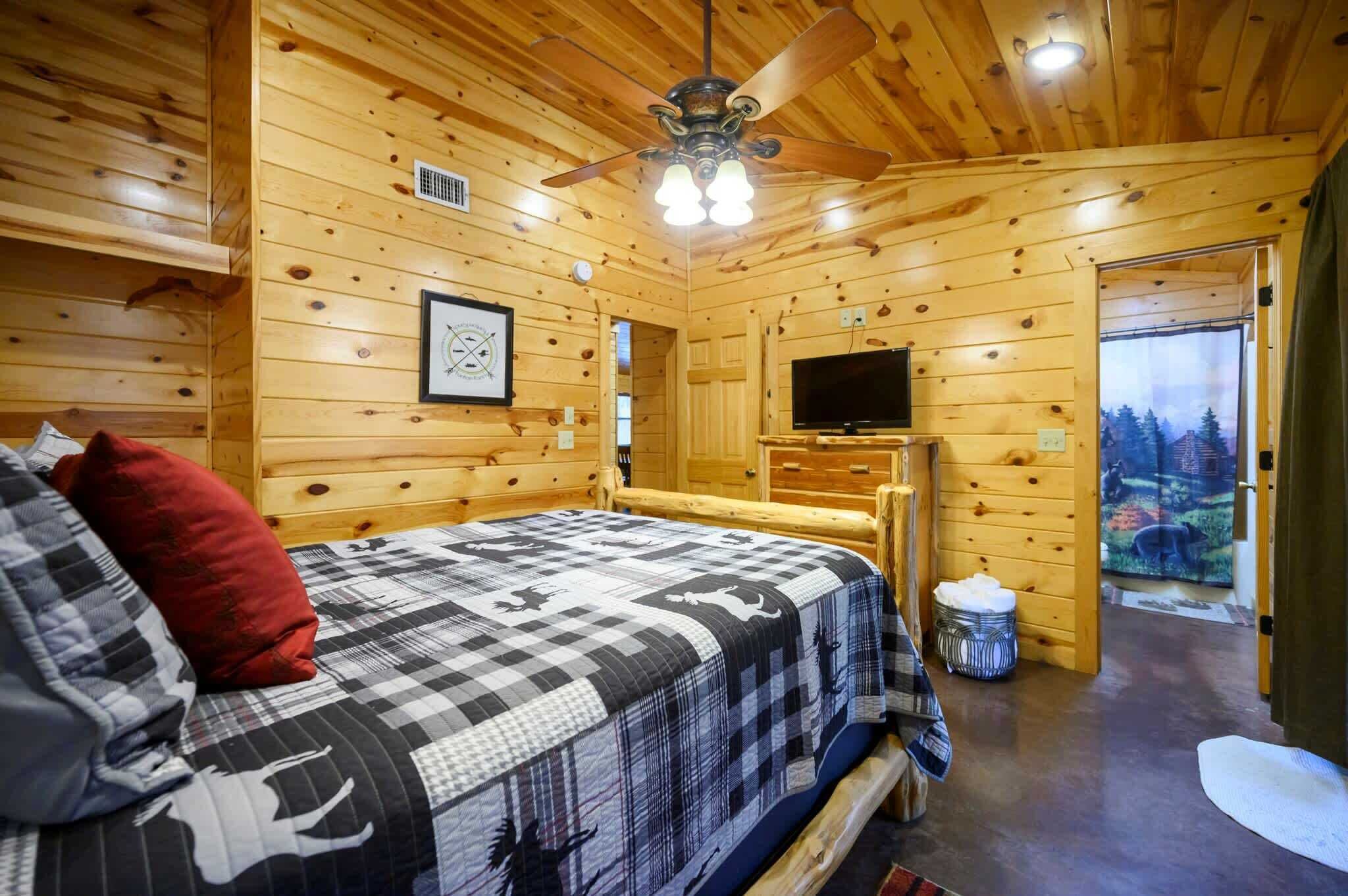 Dormitorio de cabaña de madera con cama, ventilador de techo, televisor y ventana con vista a árboles.