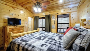 Quarto de cabana de madeira com uma cama, televisão, ventoinha de teto e janela com cortinas.