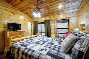 Un dormitorio tipo cabaña de madera con cama, televisor, ventilador de techo y ventana con cortinas.