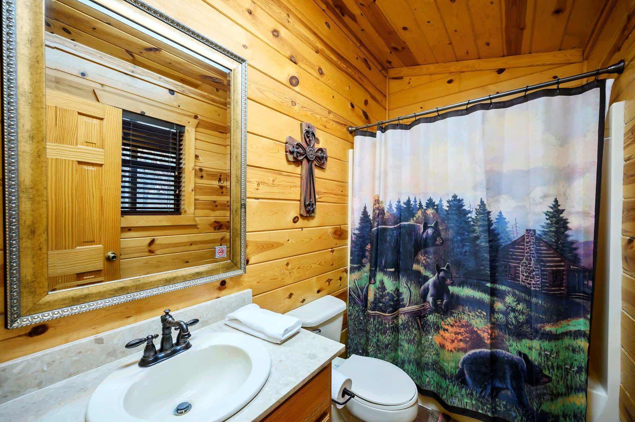 Een houten badkamer met een houten wastafel, een wastafel, een toilet en een douchegordijn met een boslandschap.