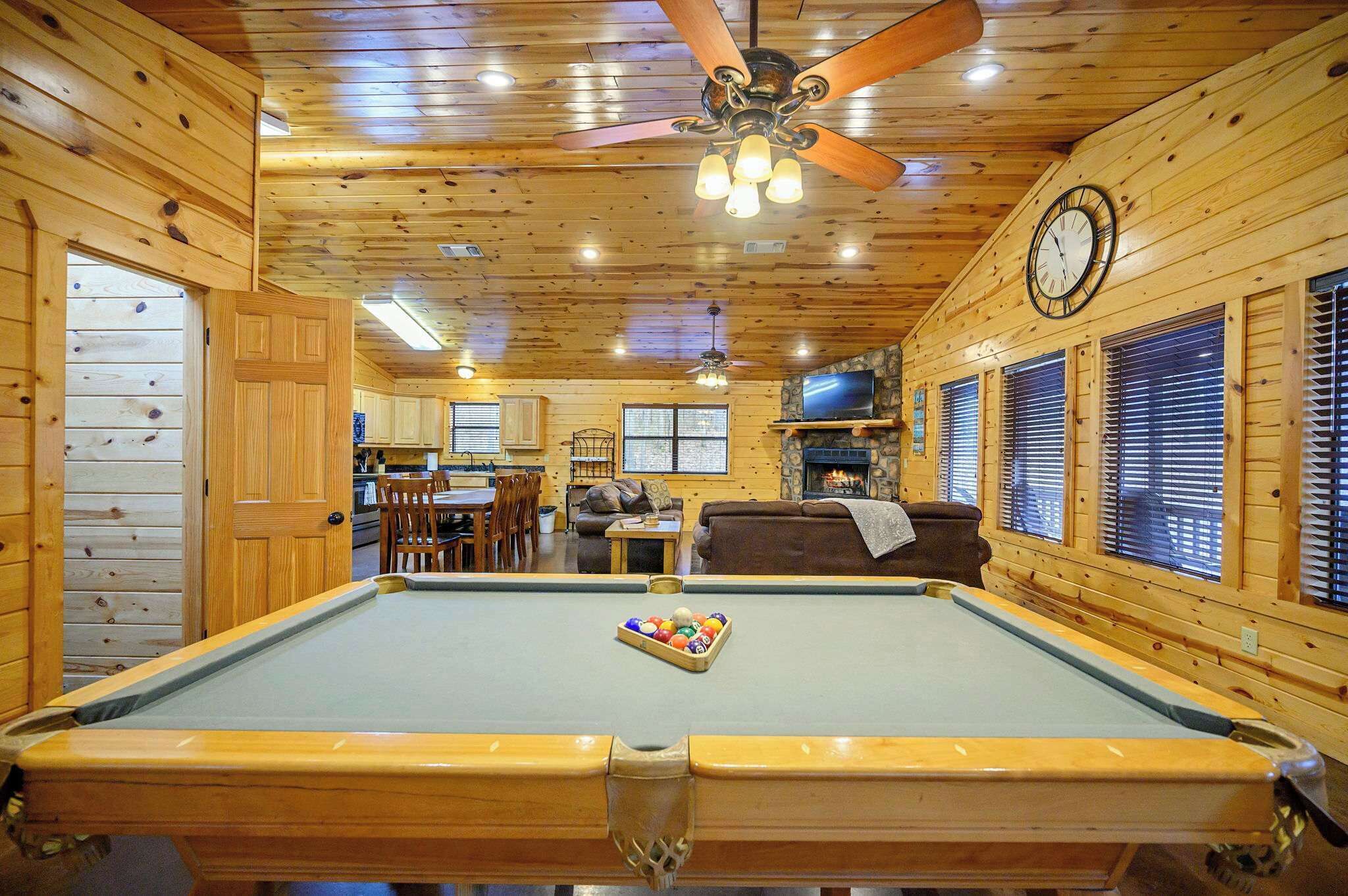 A pool table in a wooden cabin with a ceiling fan, a clock, and a fireplace.