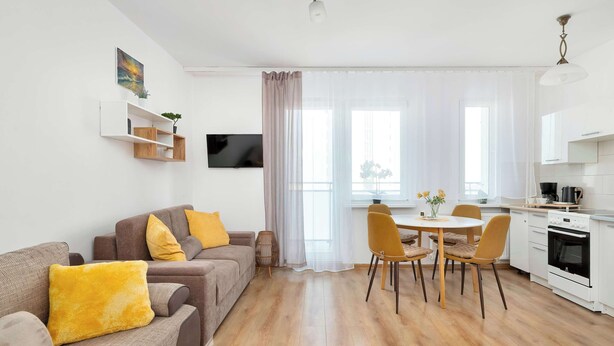 A modern living room with a sofa, a dining table with chairs, and a kitchen area with a stove and cabinets. - Sunny Studio with Sea View | Balcony (Gdynia)