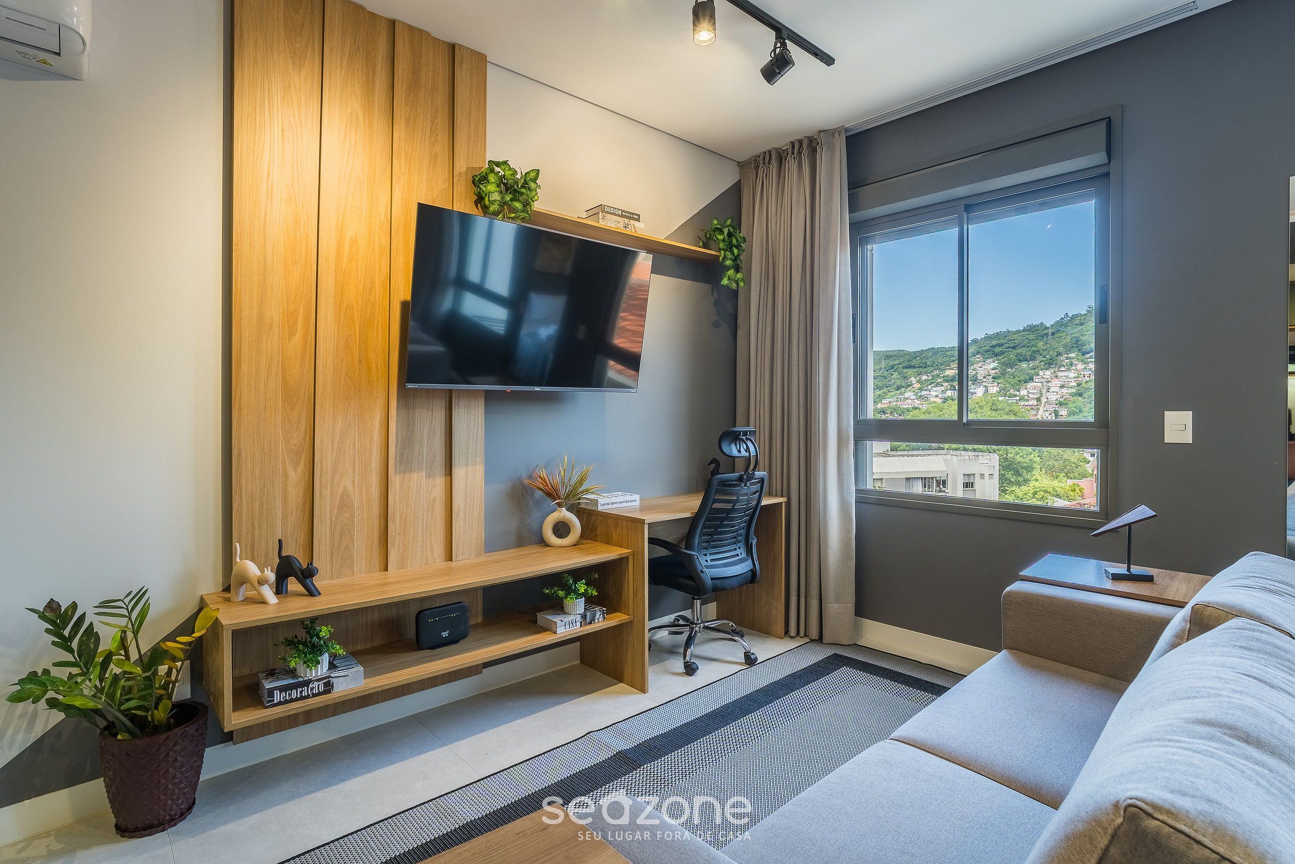 A modern living room with a flat-screen TV mounted on a wooden wall, a black office chair, a beige sofa, and a large window offering a city view.
