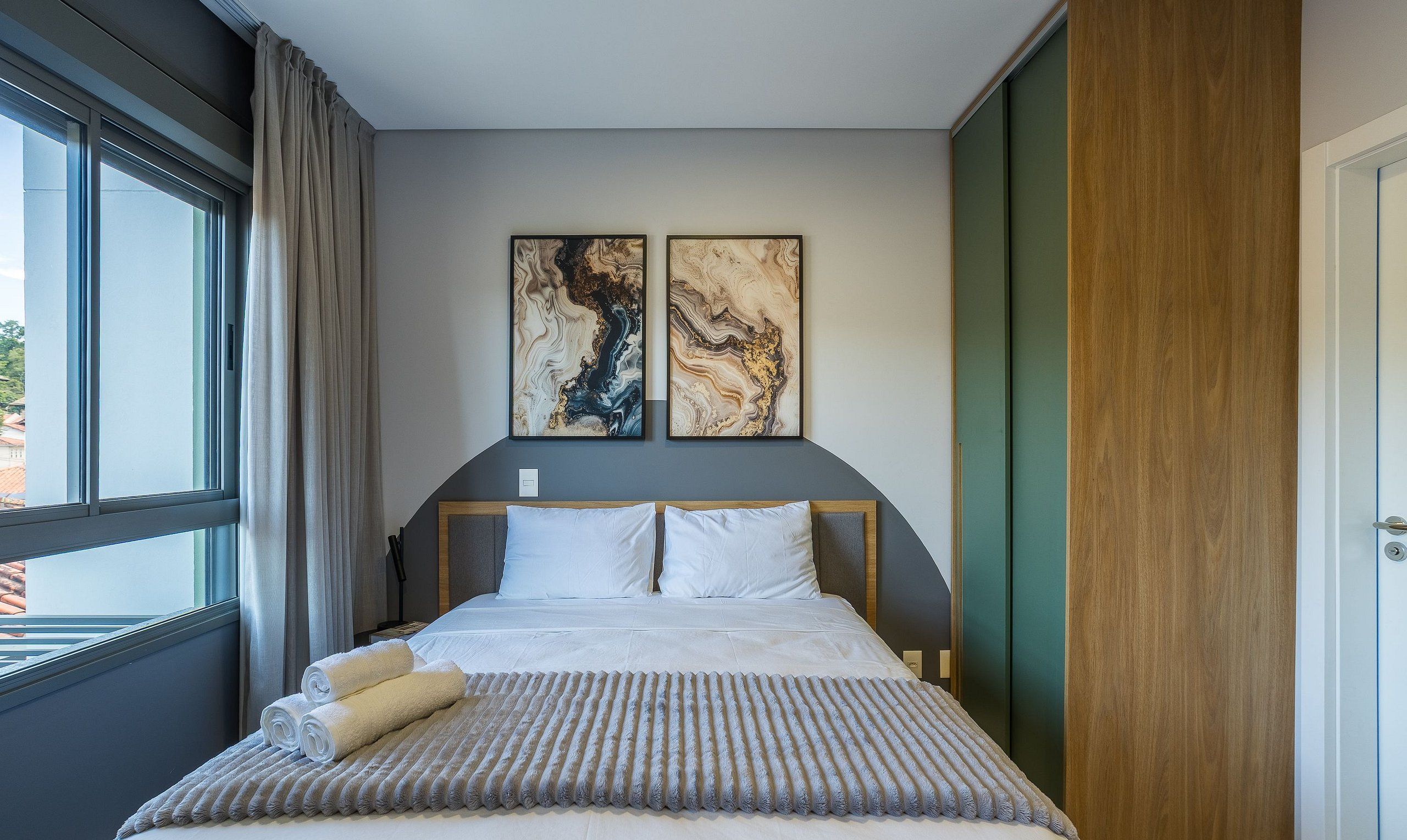 A hotel room with a bed, two paintings on the wall, a window with curtains, and a door.