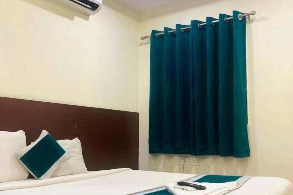 Ein Hotelzimmer mit einem Bett, weißer Bettwäsche, einer türkisblauen Decke, einem dunklen Kopfteil, türkisgrünen Vorhängen und einer Klimaanlage.